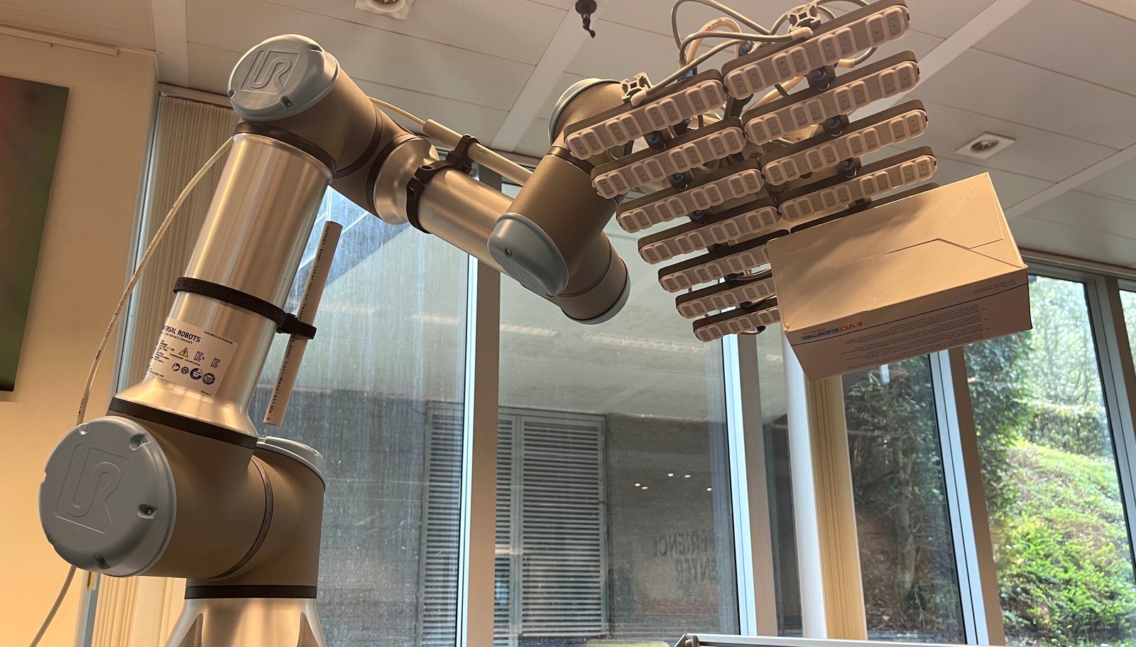 Bras robotique équipé d'une pince souple manipulant délicatement une boîte en carton dans un laboratoire lumineux de la VUB à Bruxelles