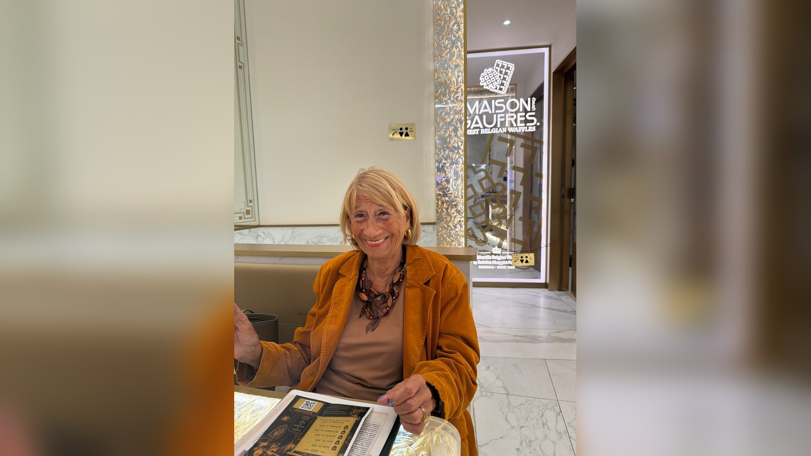 Sonia Neefs à la Maison des Gaufres (Antoine Akayyan Family, Bruxelles depuis 1982). Sonia Neefs smiling in an ochre jacket at Maison des Gaufres in Brussels