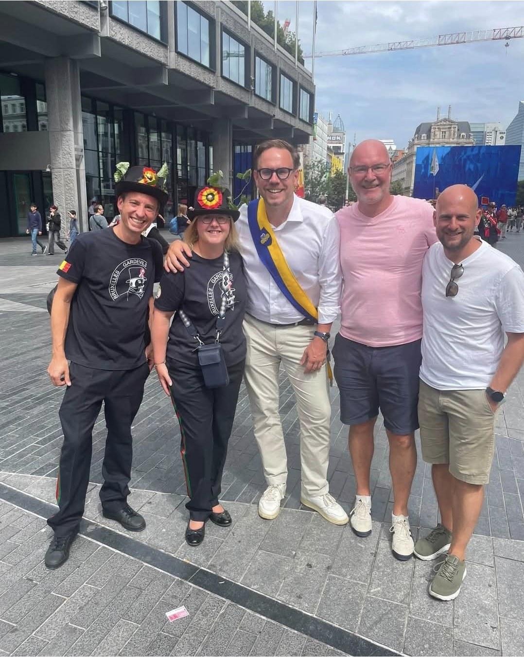 Cinq personnes posent en souriant sur une place de Bruxelles après la plantation du Meyboom : deux membres des Bruxelles Gardevils en t-shirt noir et chapeau fleuri, un élu portant l’écharpe bleue et jaune de la Ville de Bruxelles, et deux accompagnants.