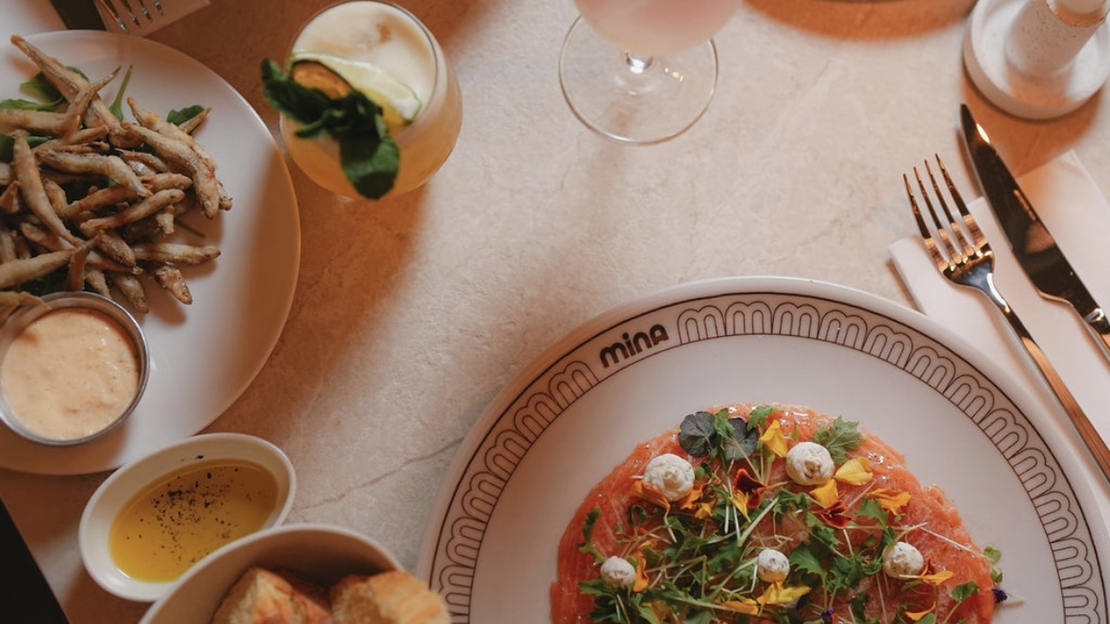 Table dressée chez Mina à Ixelles : carpaccio, fritures, pain et cocktail sur marbre clair.